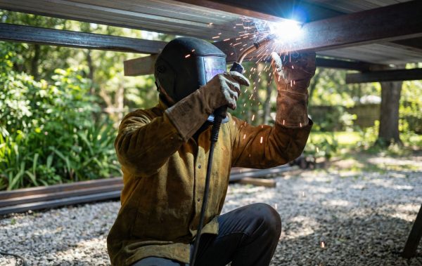 Worker wearing protective gear welding metal beams outdoors with sparks visible.